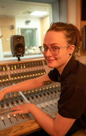 A student in a black polo shirt adjusting the controls on a recording studio mixing desk while smiling at the camera. A student in a black polo shirt adjusting the controls on a recording studio mixing desk while smiling at the camera.